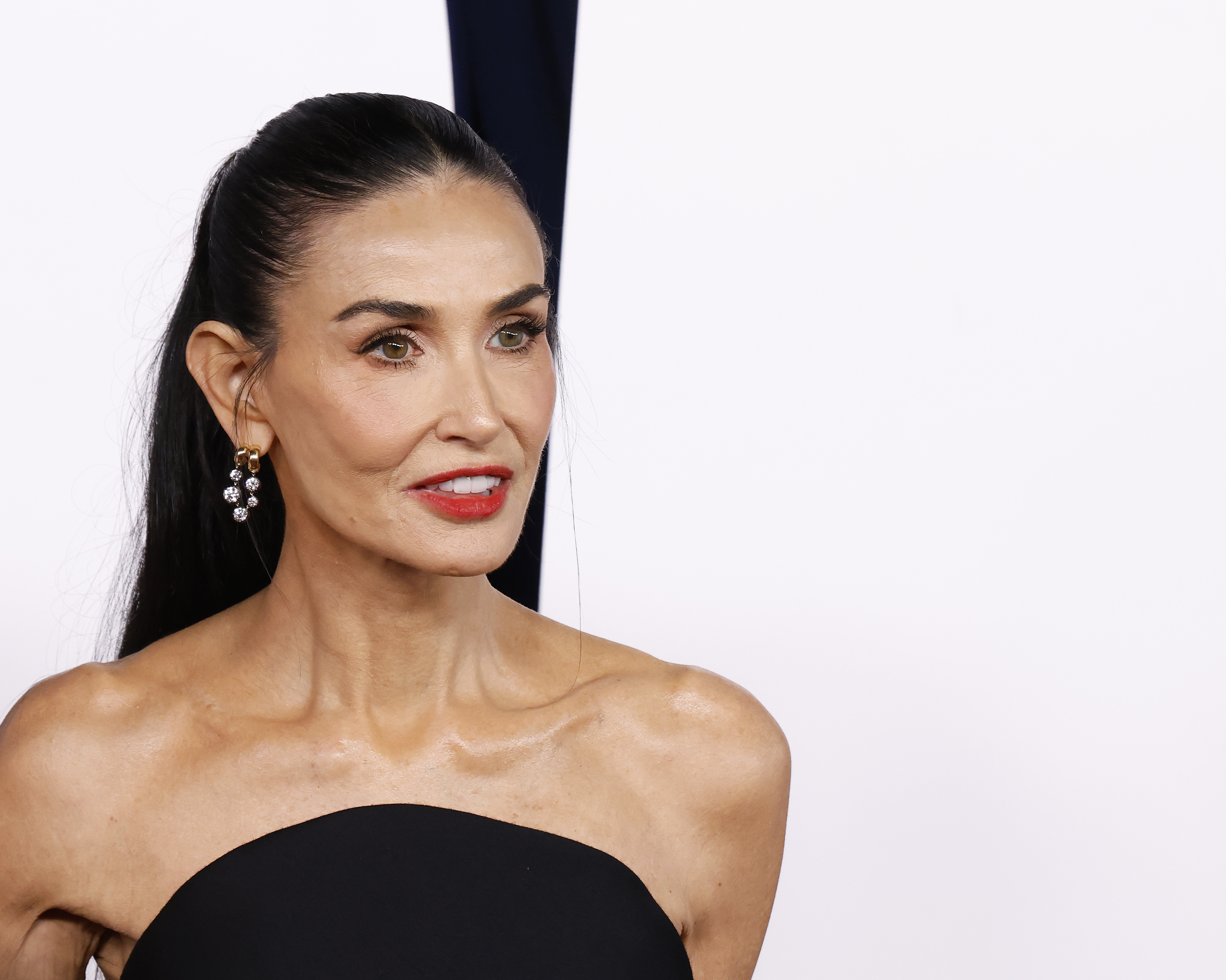 A white woman with black hair pulled back into a long ponytail. She is wearing a strapless, black gown with dangly earrings and red lipstick and posing on a red carpet in front of a white background.