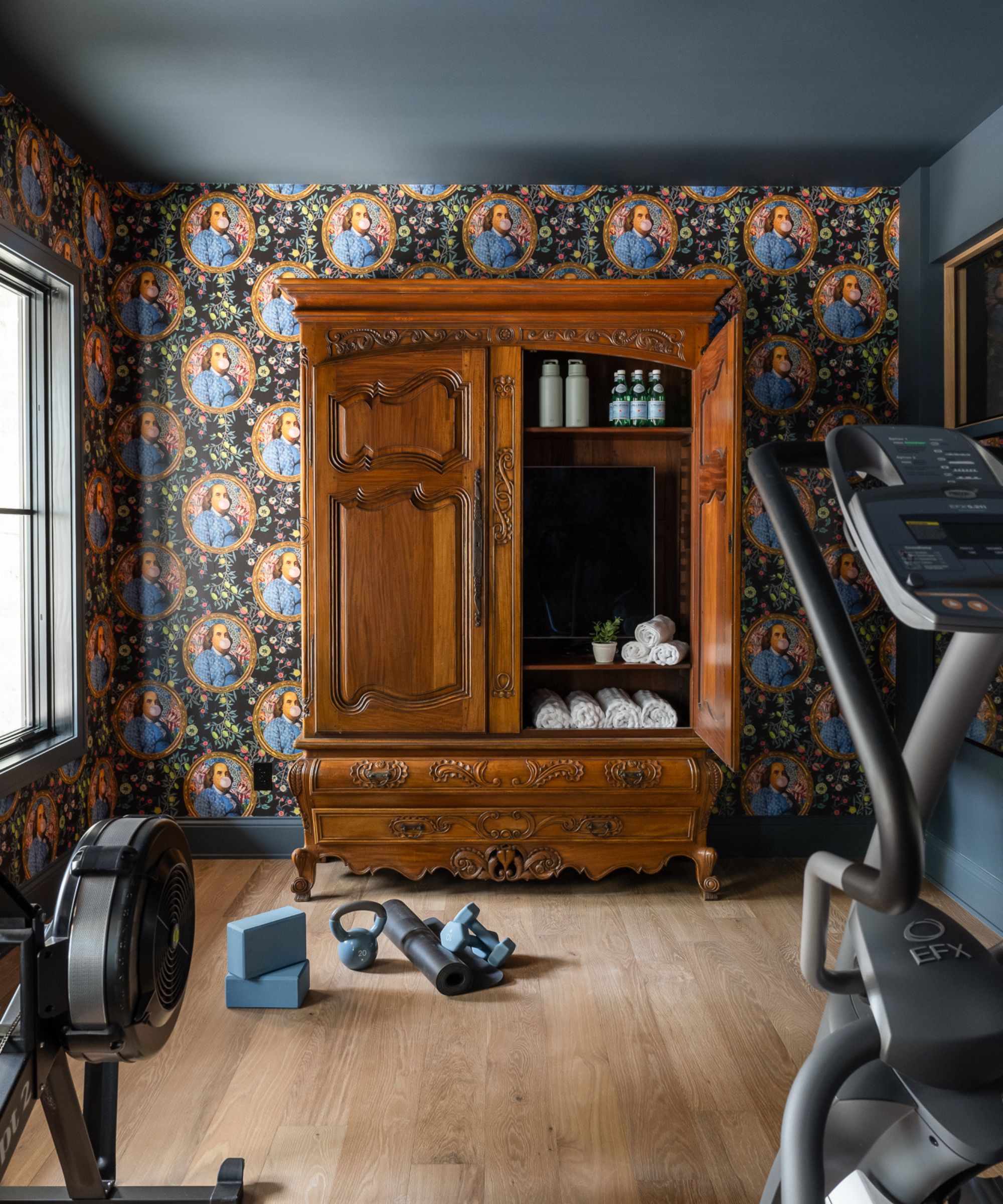 A unique home gym featuring whimsical Benjamin Franklin portrait wallpaper, a large antique wooden armoire used for storage, a rowing machine, and an elliptical.