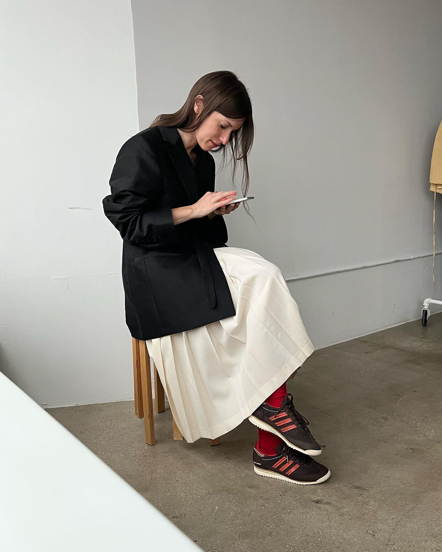 a stylish women sits on her a stool scrolling on her cellphone wearing a black blazer, pleated ivory skirt, red socks, and brown Adidas SL72 sneakers
