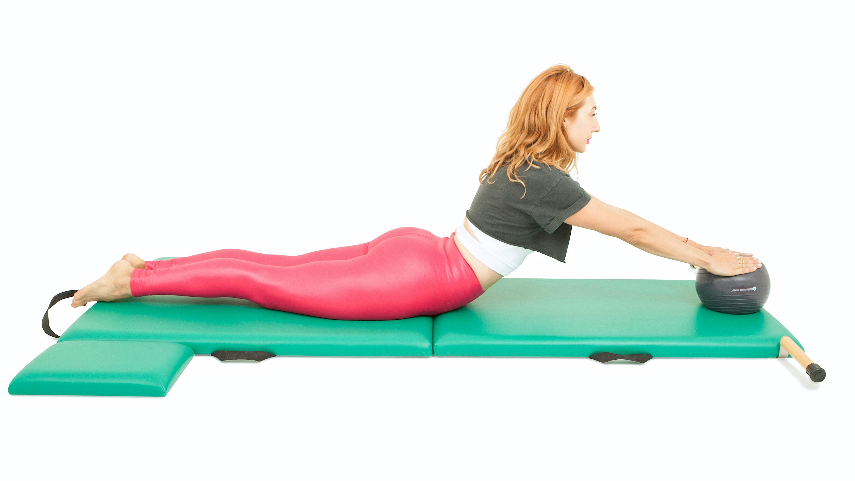 Lesley Logan performs Swan with a Pilates ball on a padded mat. She is lying on her front, her legs extended behind, her torso elevated, arms long in front and hands resting on a ball.