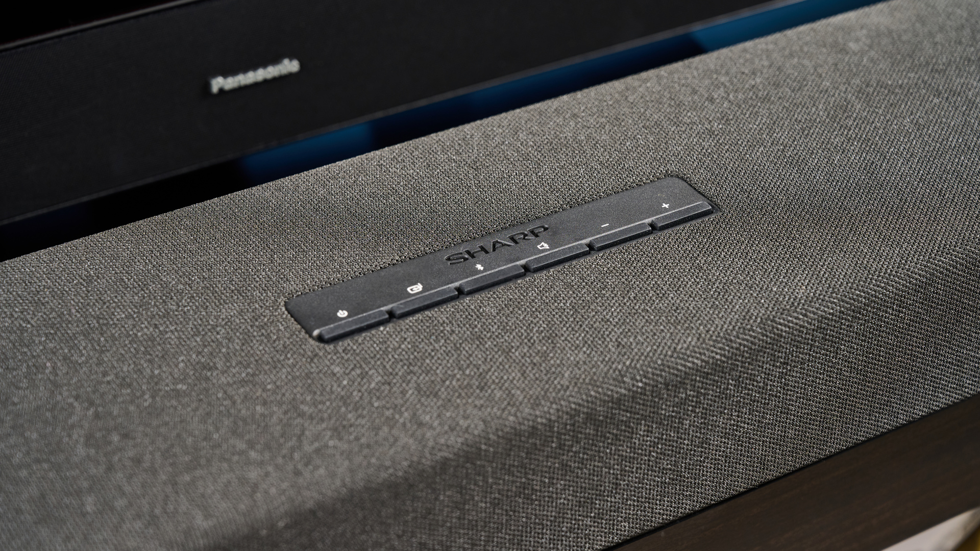 the Sharp 3.1.2 HT-SBW53121 Soundbar in black with a remote, subwoofer, against a blue background next to a panasonic TV
