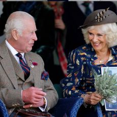 King Charles wearing a tweed jacket and vest smiling at Queen Camilla wearing a blue feather print dress and holding a sprig of greens