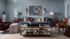 Pale blue living room with blue sofa, glass mirrored coffee table, antique chairs and tables, crown moulding and panelling on the wall, and eclectic decorations.