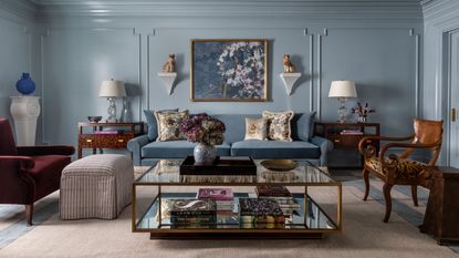 Pale blue living room with blue sofa, glass mirrored coffee table, antique chairs and tables, crown moulding and panelling on the wall, and eclectic decorations.