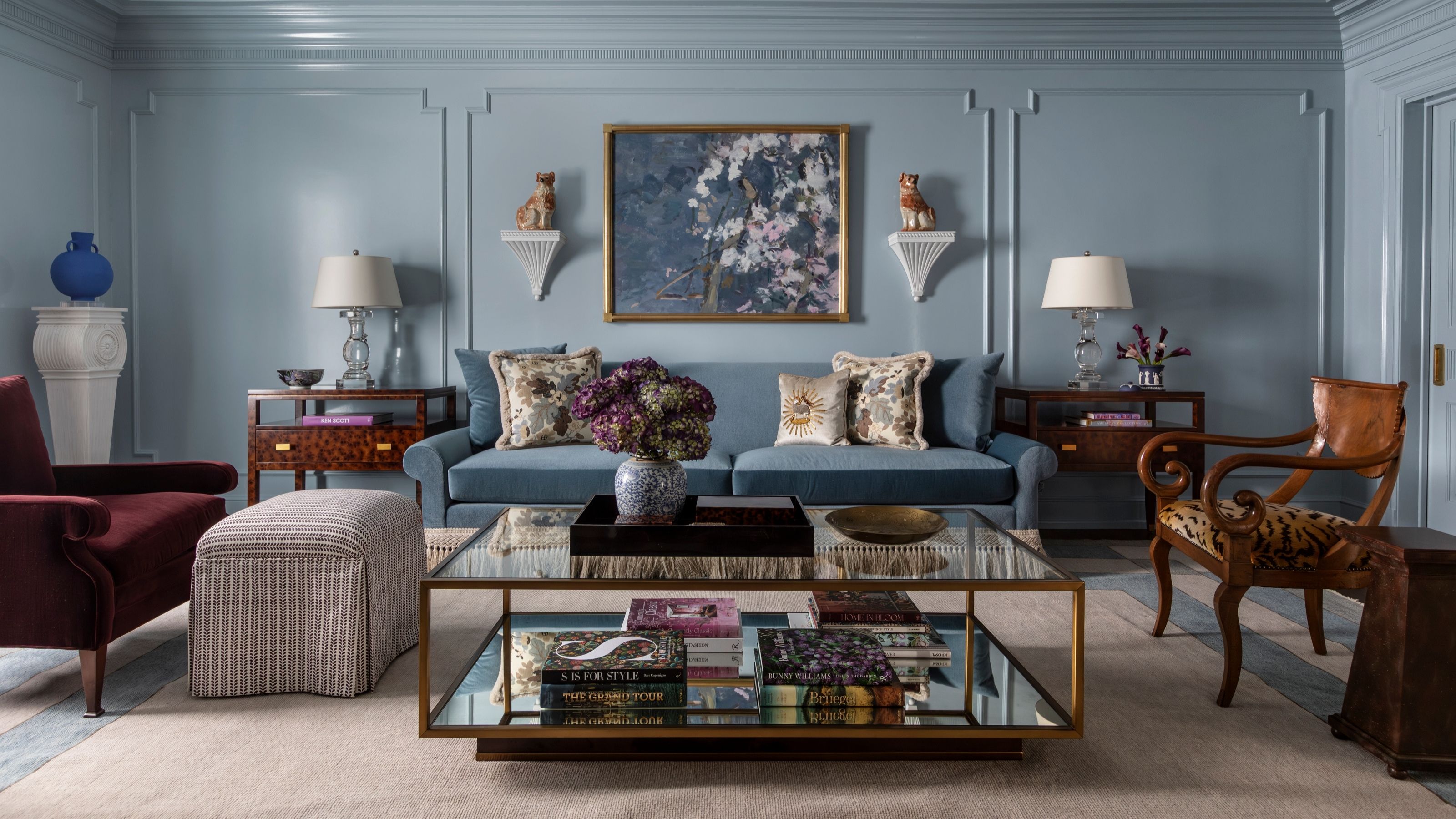 Pale blue living room with blue sofa, glass mirrored coffee table, antique chairs and tables, crown moulding and panelling on the wall, and eclectic decorations.