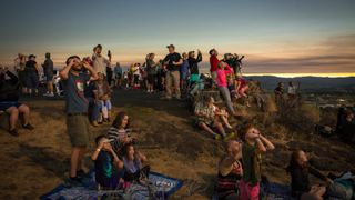 A group of people on a hillside wearing eclipse glasses are looking up at the sky, 