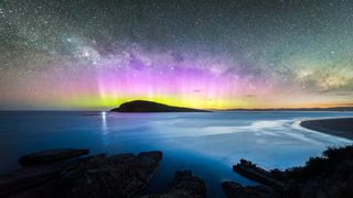 An island in the distance is silhouetted against a starry sky filled with aurora, a yellow band of light lies closer to the horizon and a bright pink aurora appears above.