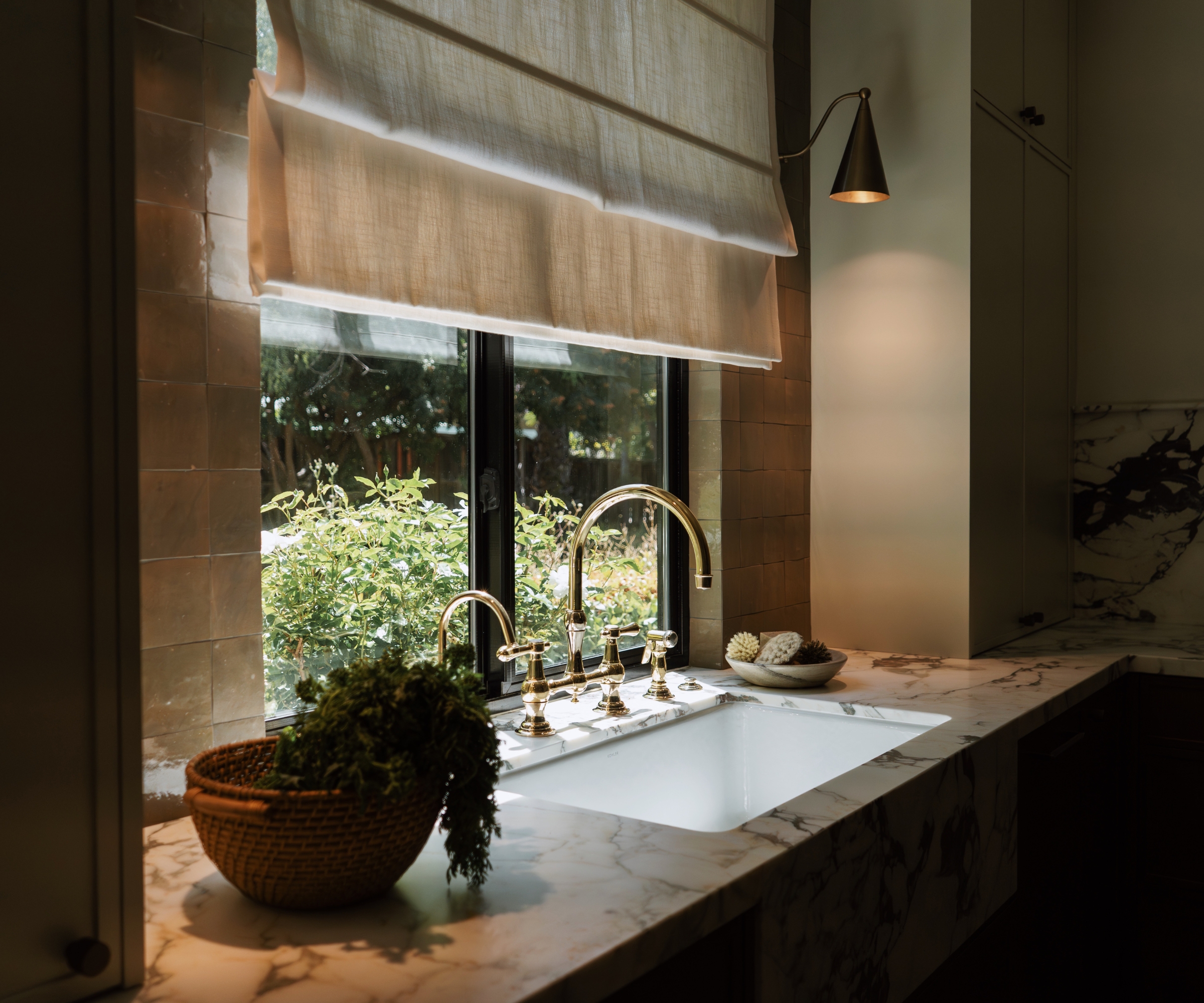 The kitchen sink below a window covered in a linen blind and flanked by two wall sconces with warm-toned bulbs