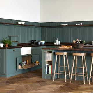 Green kitchen with panelling halfway up the wall, white paint the remainder of the wall, matching green panelled kitchen island with wooden bar stools, and dark wooden floorboards