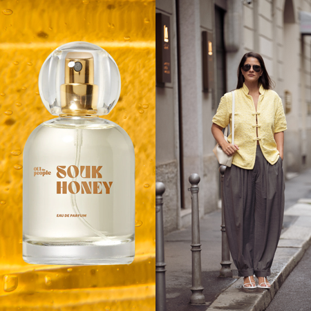 A woman wearing a yellow top and grey pants next to a bottle of honey perfume. 
