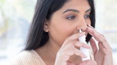 A close up of a woman with straight black hair wearing a pink and white striped shirt who holds a canister of nasal spray to her right nostril