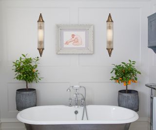 two brass and glass wall lights either side of framed print above roll top bath with two grey plant pots and small orange trees either side