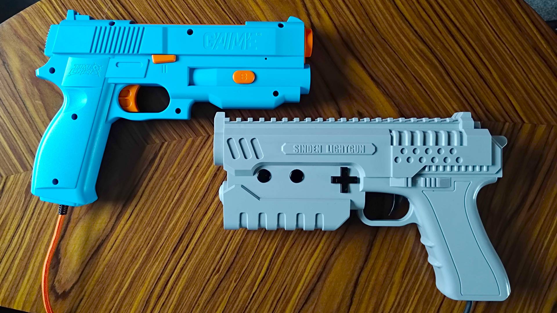 G&#039;AIM&#039;E Time Crisis lightgun next to Sinden Lightgun on woodgrain table.