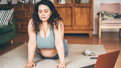 A woman practices a yoga cobra pose in her home. The lower half of her body, from her hips down, is on the floor, while her torso is erect, held up by her arms, which are straight and pushing against the ground. Her eyes are closed; in front of her is a laptop, next to her is a pair of headphones and a mobile, and behind her is a chest of drawers and arm chair. 