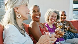 Four happy woman drinking wine together whilst sitting in a line on an urban rooftop.