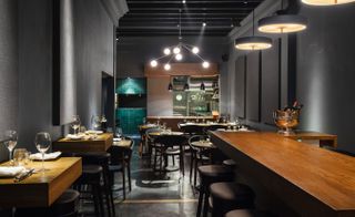 Interior view of the dining area at Cure featuring dark grey walls, two different styles of pendant lights, wooden dining tables with tableware and dark coloured chairs. There is also a partial view of the kitchen