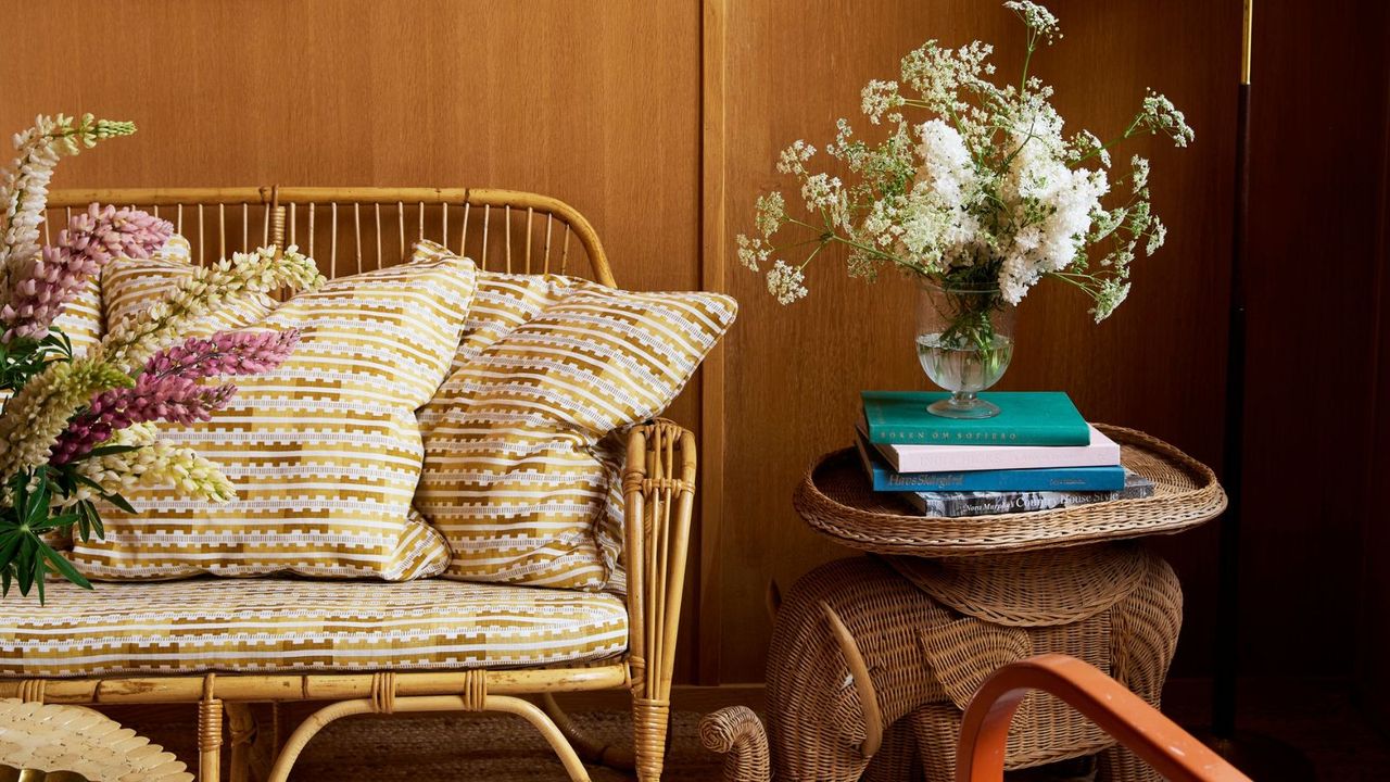 A woven sofa next to a wicker side table, with a pile of books and fired flowers in a vase on top. 