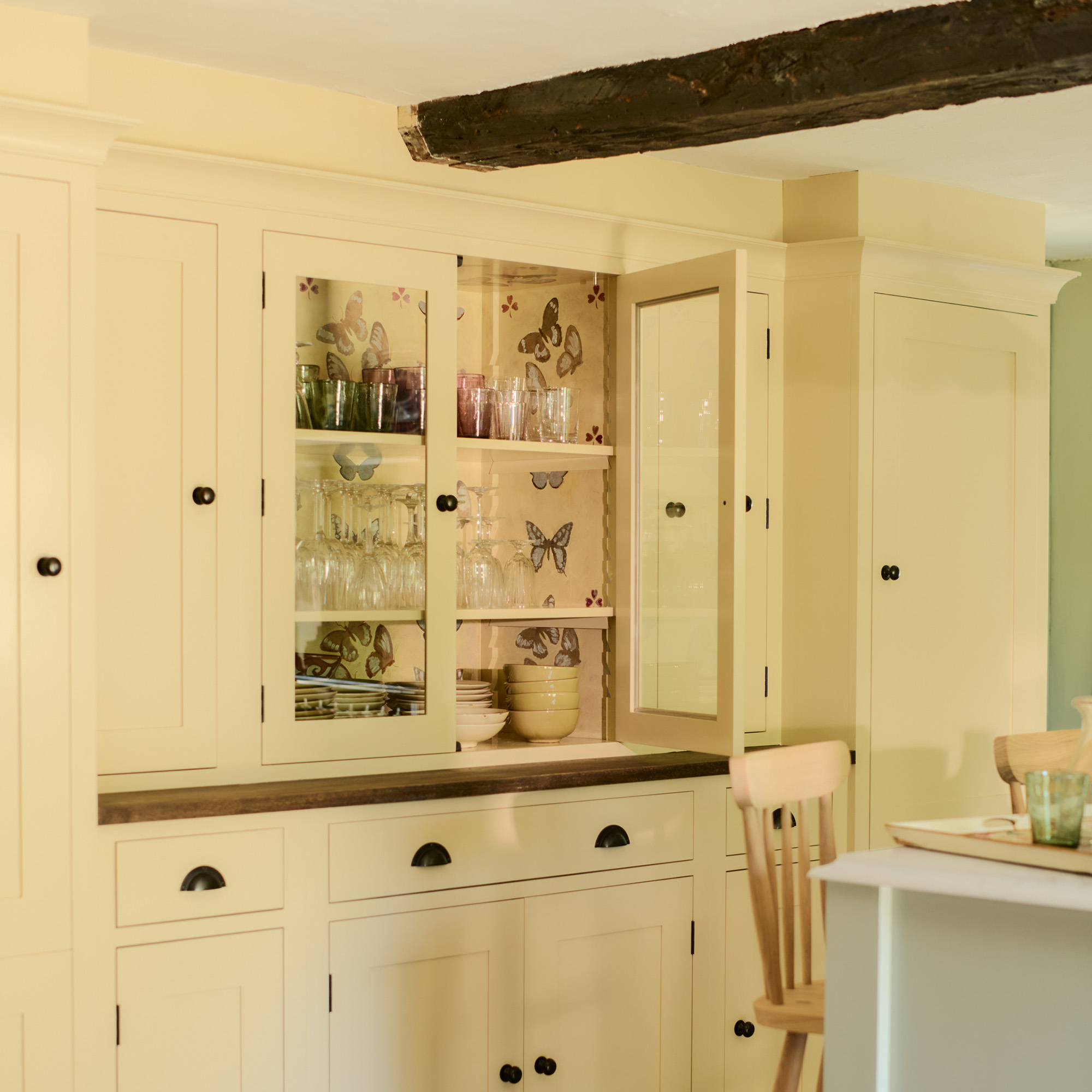 fitted pantry cupboard in cream with wooden worktop and dark handles a central glass fronted section reveals glassware and a handpainted butterfly interior