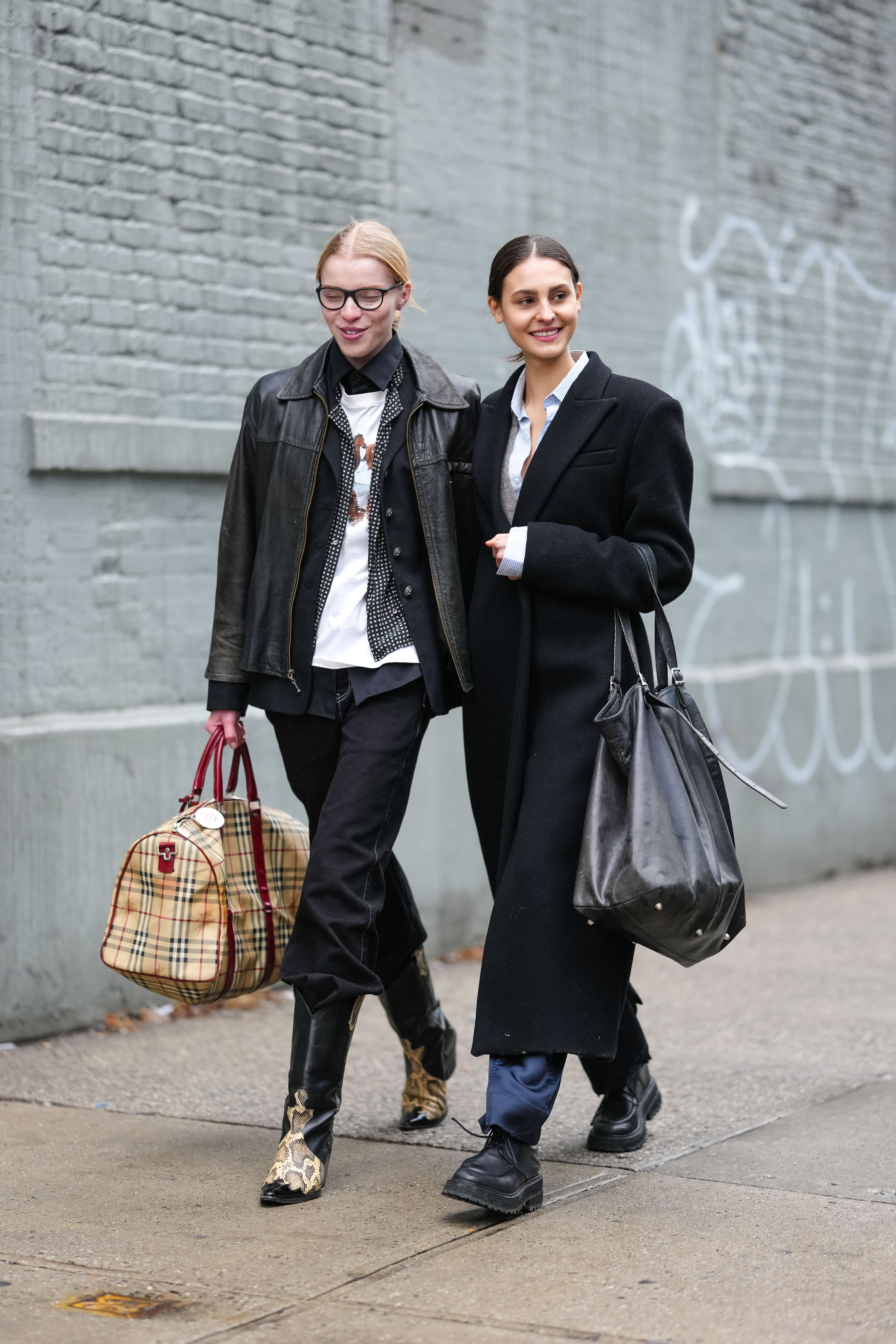 NEW YORK, NEW YORK - FEBRUARY 06: A model wears black glasses, white graphic print t-shirt, black checkered pattern/print open buttoned up long sleeve shirt, black open blazer jacket, shiny black leather jacket, light brown plaid check pattern Burberry duffle bag, black slouchy denim jean pants, shiny black pointed toe snake skin pattern/print cowboy leather boots. A model wears white buttoned up long sleeve shirt, light gray sweater, black wool coat, shiny dark blue satin loose trouser pants, shiny black oxford moccasin leather shoes, shiny black large leather bag, outside Brandon Maxwell, during the New York Fashion week Fall/Winter 2025 on February 6, 2025 in New York, New York. (Photo by Edward Berthelot/Getty Images)