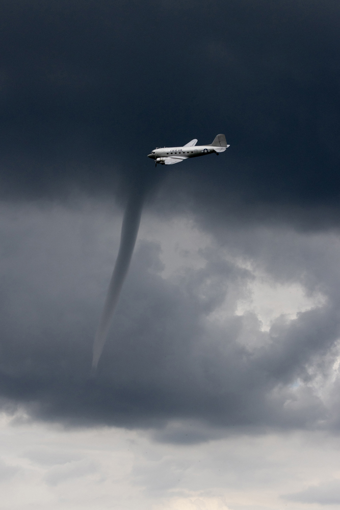A plane flies near dark storm clouds, with a visible funnel extending from the clouds, suggesting severe weather conditions