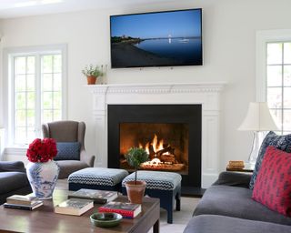 bright white living room with large black and white fireplace, grey sofas and lounge chair, twin patterned navy footstools, dark wood coffee table, built in white shelving unit, wall mounted tv above fireplace