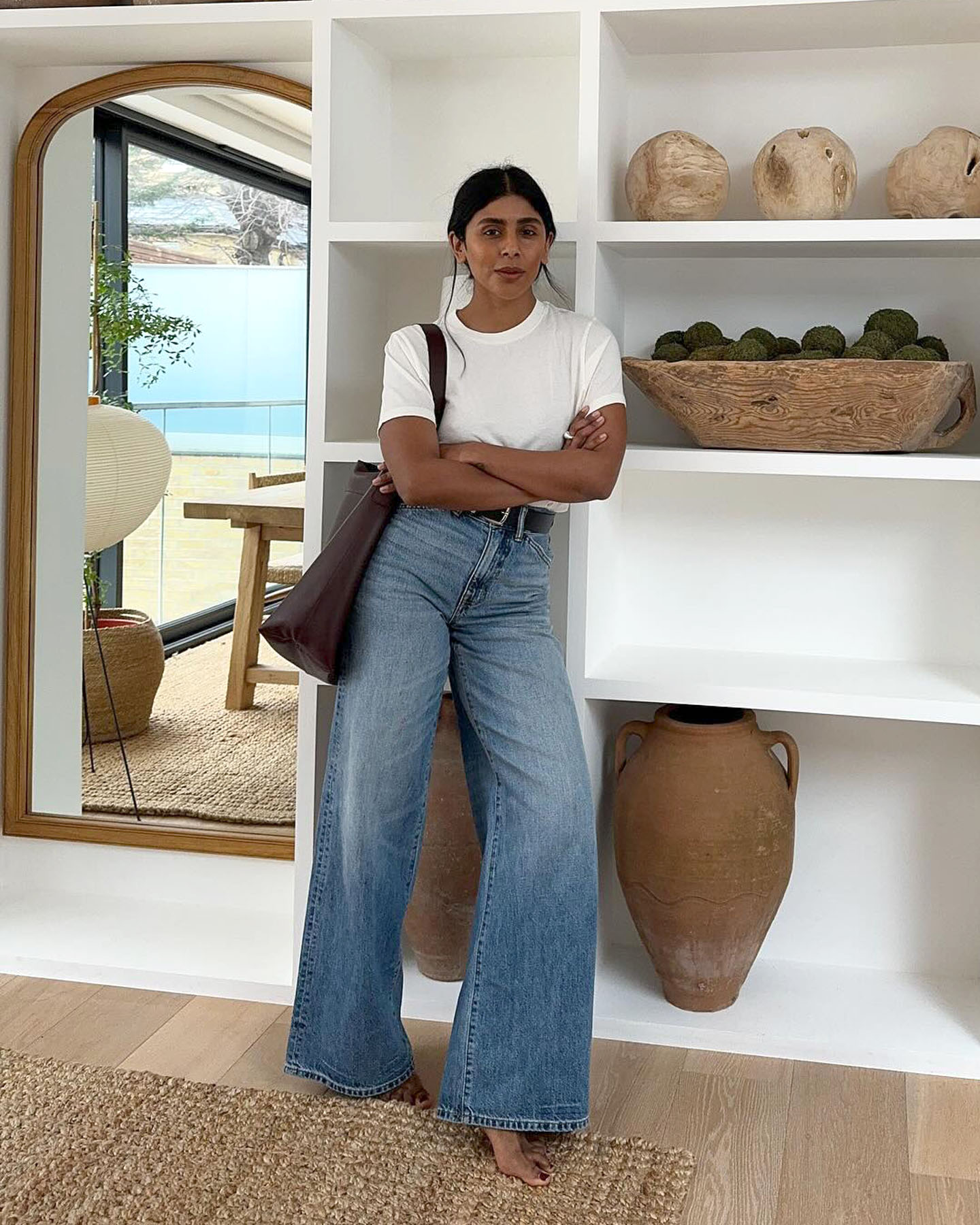 fashion influencer Monikh Dale poses in her modern London home wearing a casual outfit with a basic white t-shirt, black belt, high-waisted wide-leg jeans, and a large brown tote bag