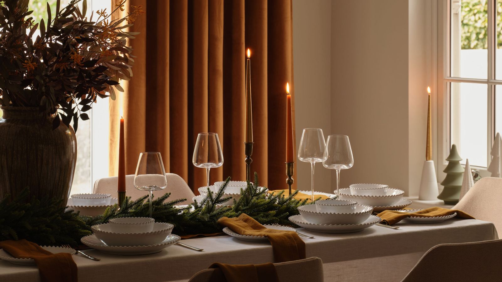 A dining table set with white bowels and decorated with a garland, candlesticks, and ceramic Christmas tree statues