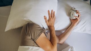 A man who is unable to sleep at night covers his head with a white pillow while holding a white alarm clock in the other hand