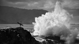 A surfer standing on a rocky outcrop, holding a surfboard, gazes at the crashing waves in a black and white landscape. The ocean stretches out in the background, while a large wave splashes dramatically against the rocks, emphasizing the power of the sea. The scene is set against a backdrop of dark mountains under a cloudy sky.