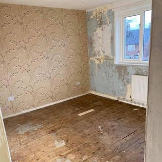 Stripped back room with exposed floor boards, wallpaper taken off one wall with a white window, and a brown flower pattern wallpaper still on another wall