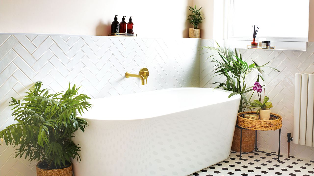 Freestanding modern white bath in a white tiled bathroom with plants in baskets