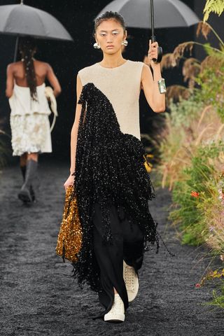 A female model wearing a long sleeveless black, brown and gold dress walking down a runway made of black sand.