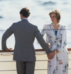 Princess Diana wearing a white floral dress standing on a ship with her back to the water while Prince Charles, wearing a gray suit, is turned the other way and holding her hand