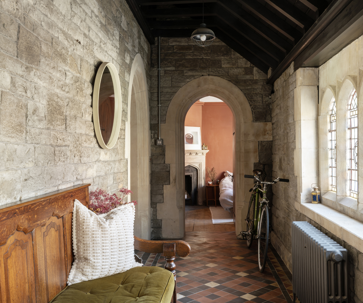 Hallway with brick walls to the left, stained glass windows to the right, and an arch door way straight ahead showing a glimpse into a peach-coloured room with fireplace