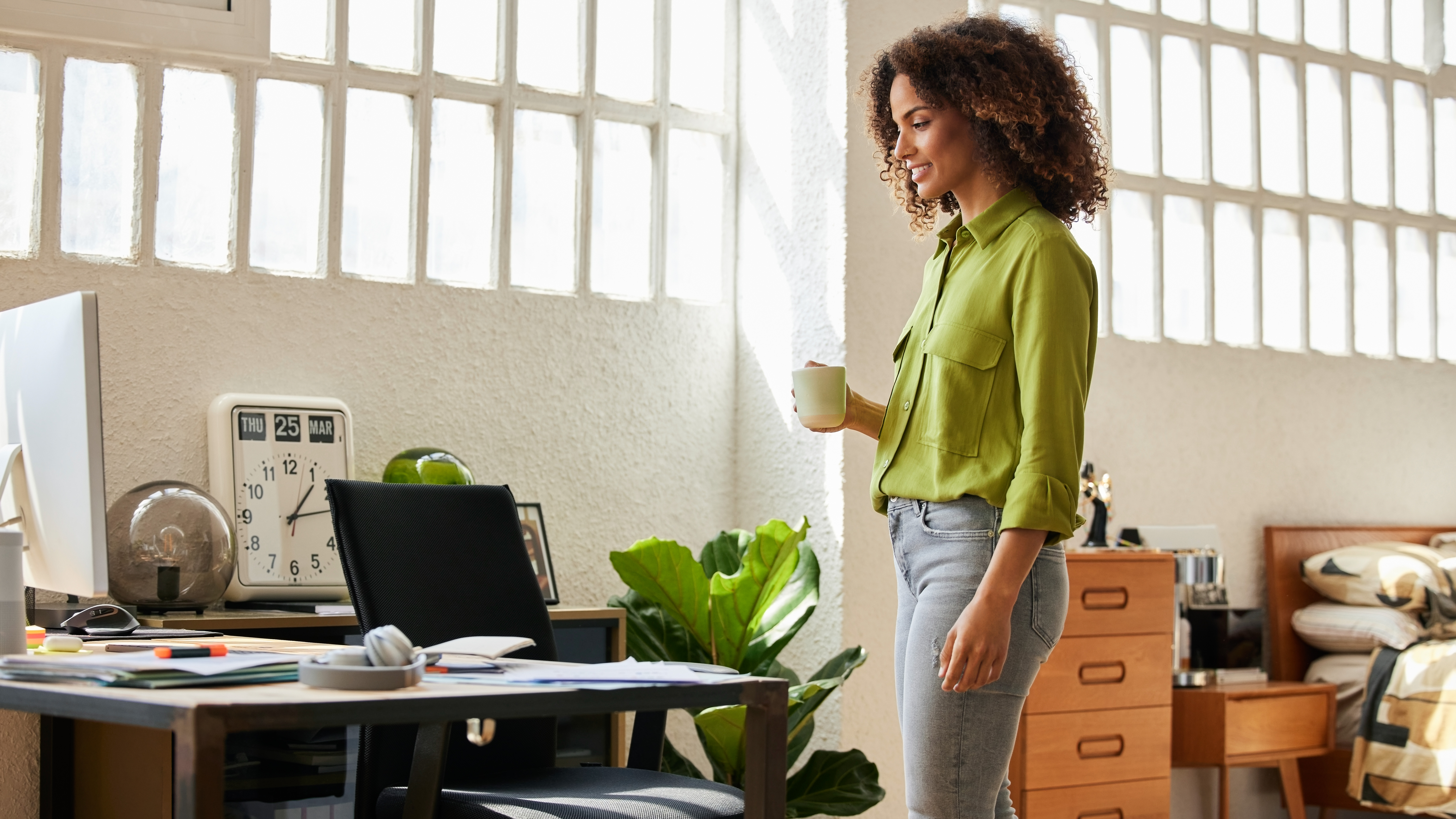 A woman walks across her office/ bedroom holding a cup of coffee. We see a messy desk with a laptop, a leafy plant, a chest of drawers and a bed in the room.