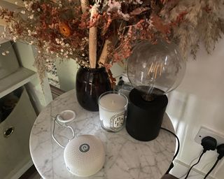 Dreamegg D11 small white noise machine in white on marble side table with Diptyque candle, vase of dried flowers and a black lamp with exposed bulb
