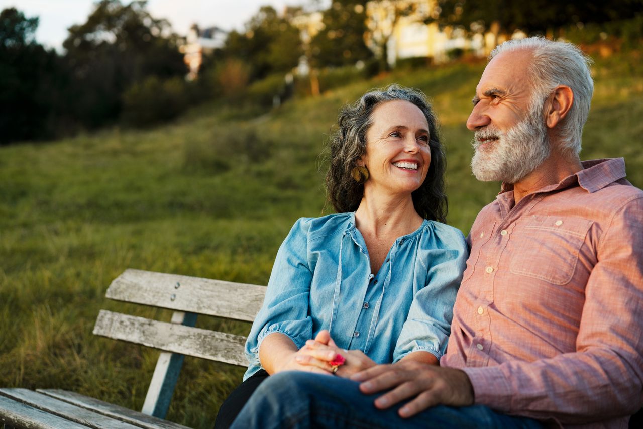 A happy couple aged approx 50-60 sat on a park bench together.