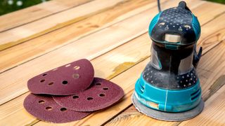 Teal and black orbital sander sat next to three circular sanding discs on a wooden table