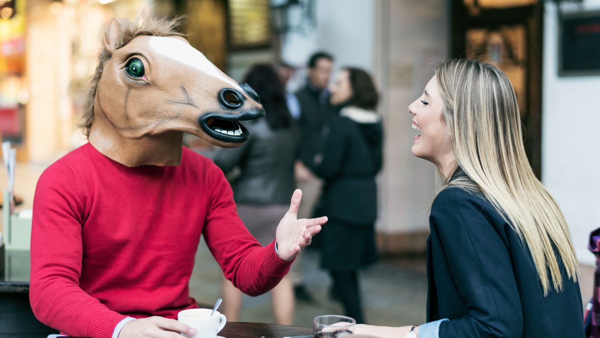 woman sat at an al fresco table laughing, talking to a man wearing a horse mask