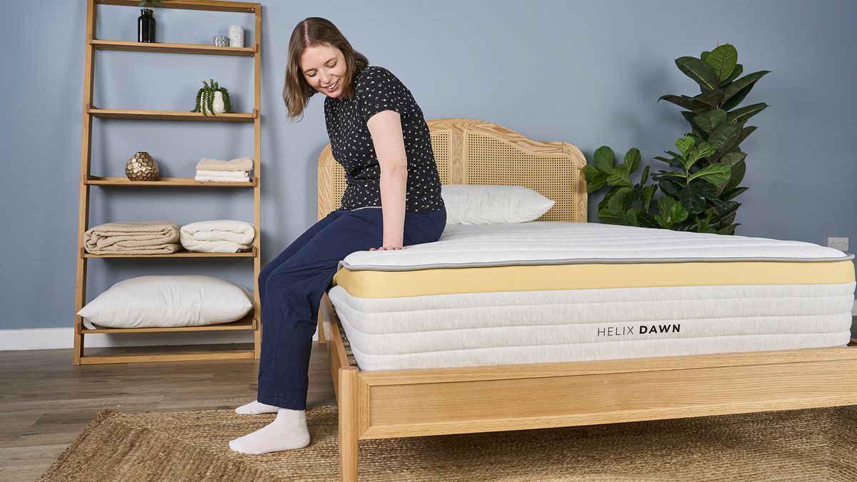 A woman sits on the side of the Helix Dawn mattress on a bed frame in a bedroom, testing the edge support