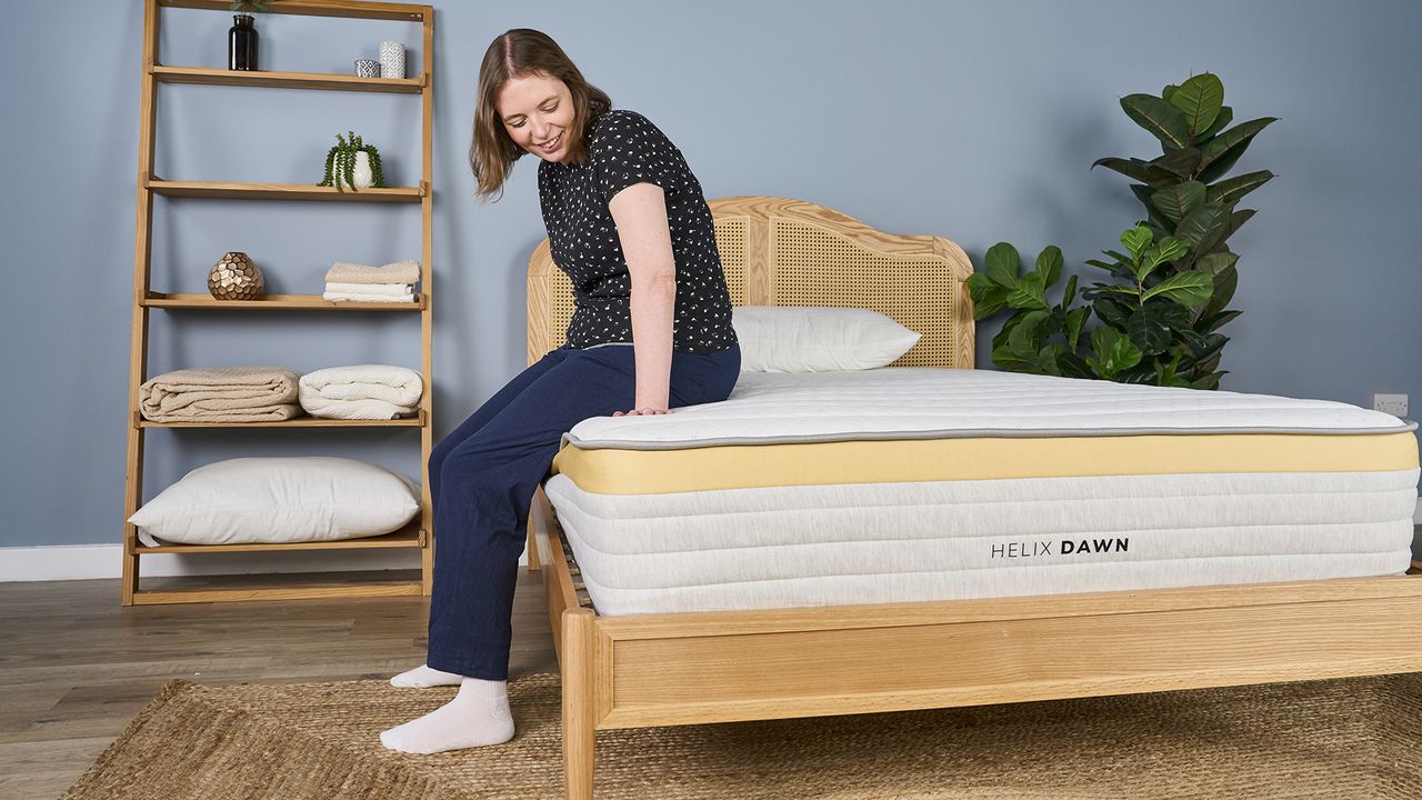 A woman sits on the side of the Helix Dawn mattress on a bed frame in a bedroom, testing the edge support