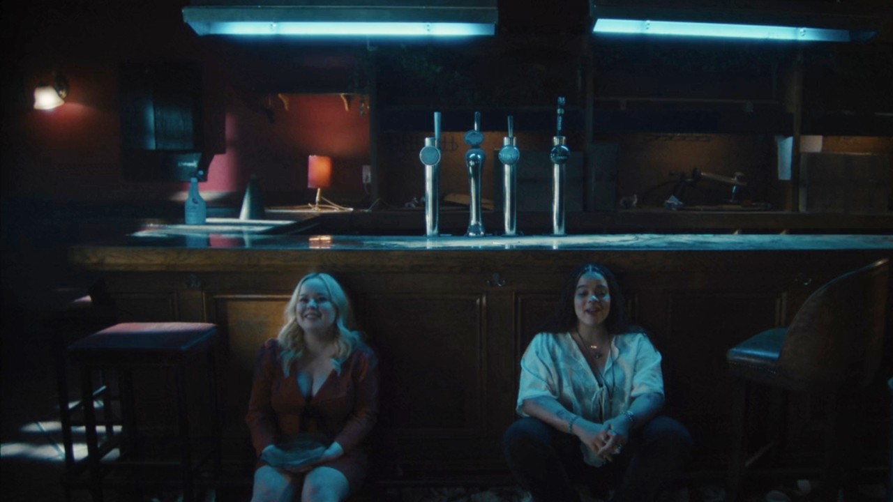 Nicola Coughlan and Lydia West sitting on the floor in the bar in Big Mood. 