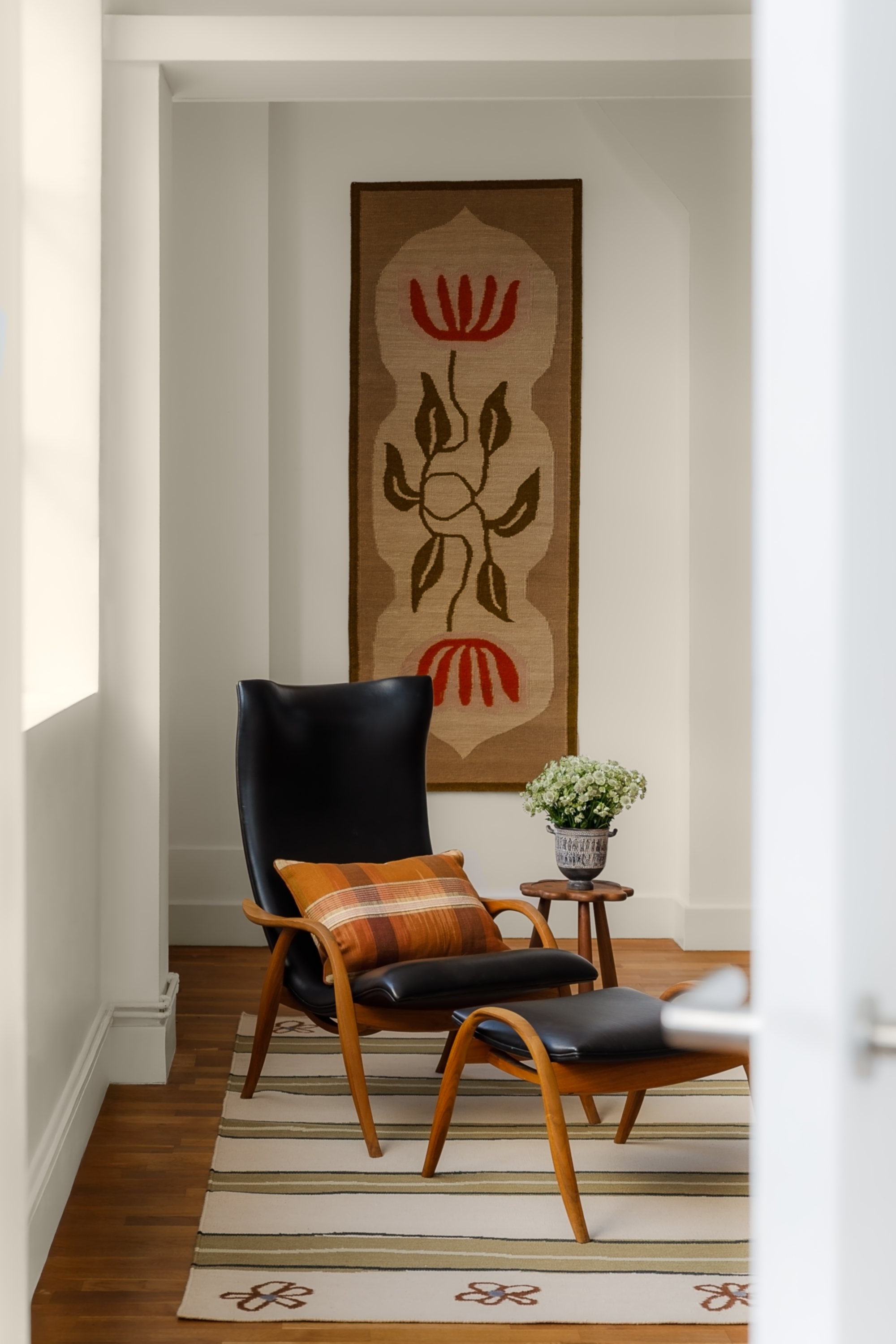 Image of a white room with a green and cream striped rug on the wooden floor with a black leather and wooden armchair and a matching stool on top of it. There is a brown rug with a red flower on it hanging on the wall.