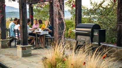 wood pellet grill on a patio with people sat around a dining table underneath a pergola