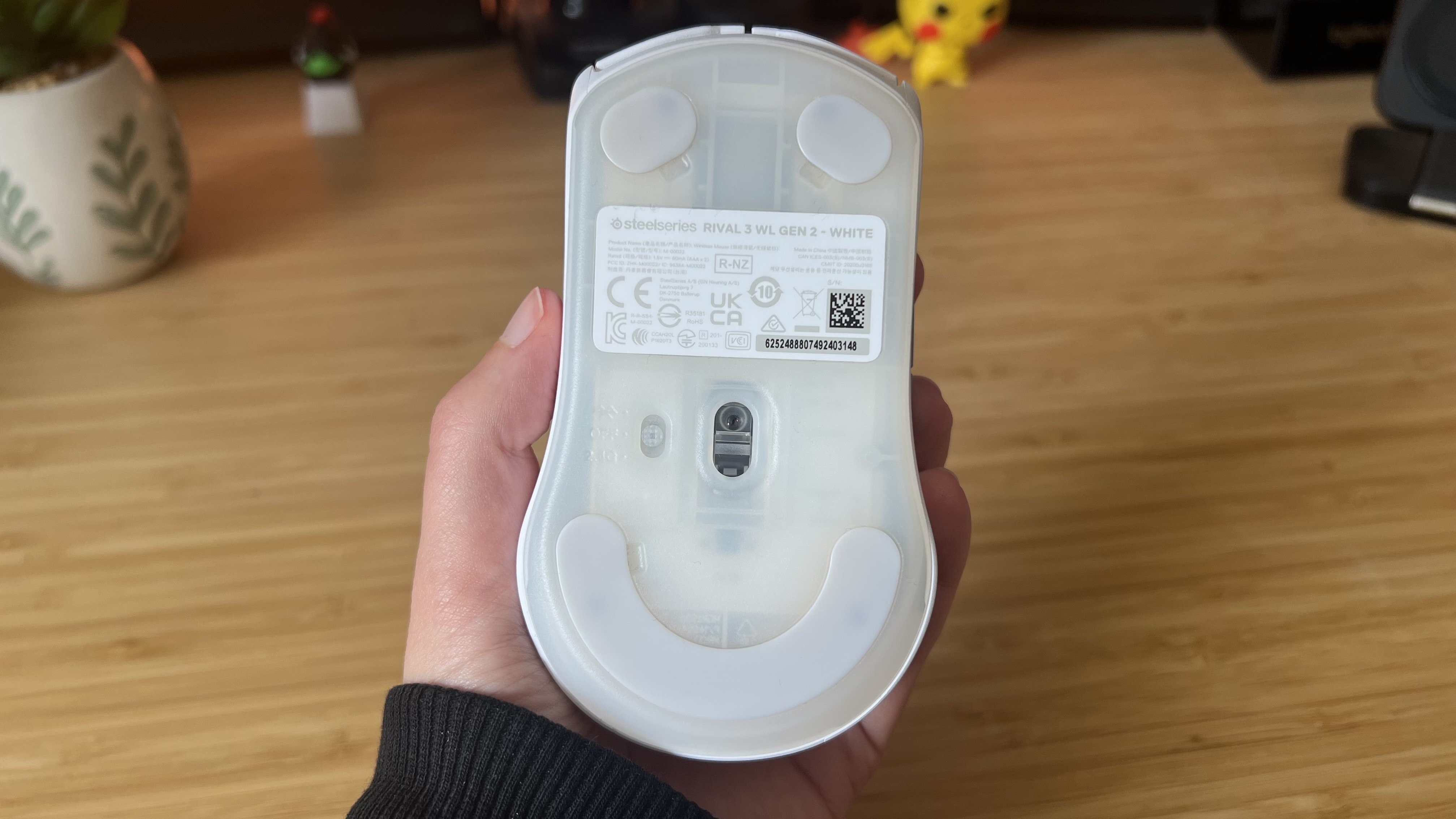 Hand holding SteelSeries Rival 3 Wireless Gen 2 gaming mouse upside down to show sensor and feet underneath