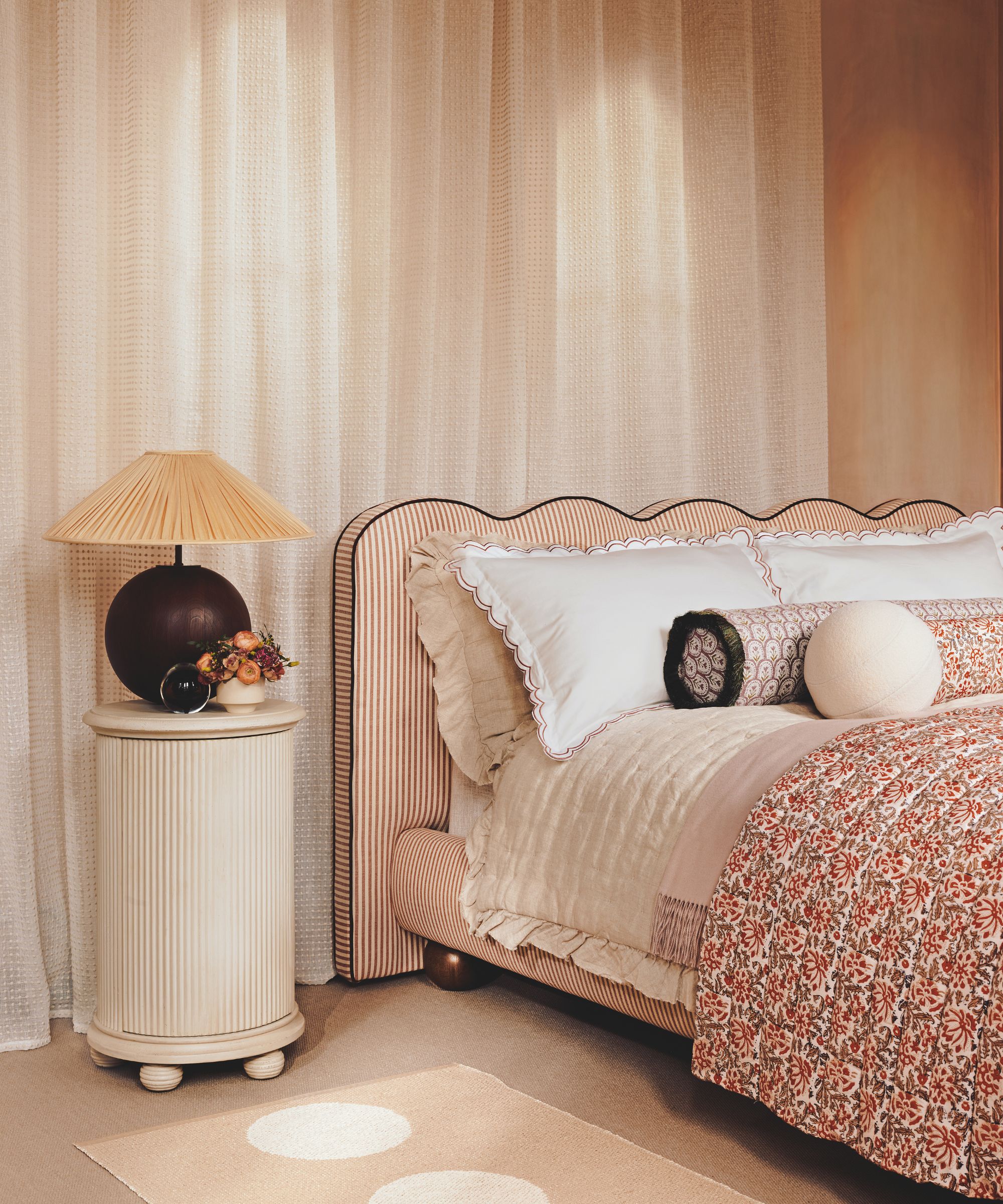A dreamy bedroom setup featuring a bed with a scalloped, striped headboard and floral linens. A fluted white nightstand holds a dark spherical lamp with a pleated tan shade against sheer, floor-to-ceiling curtains.