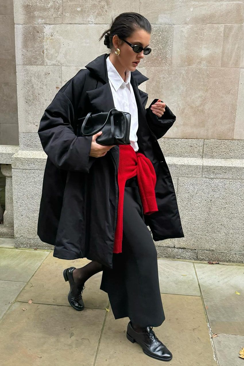 A woman wearing a black coat, a white button-down shirt, black pants, and black derby shoes