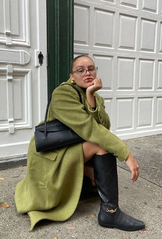 A woman wearing wearing a long green coat with a black handbag and black riding boots.