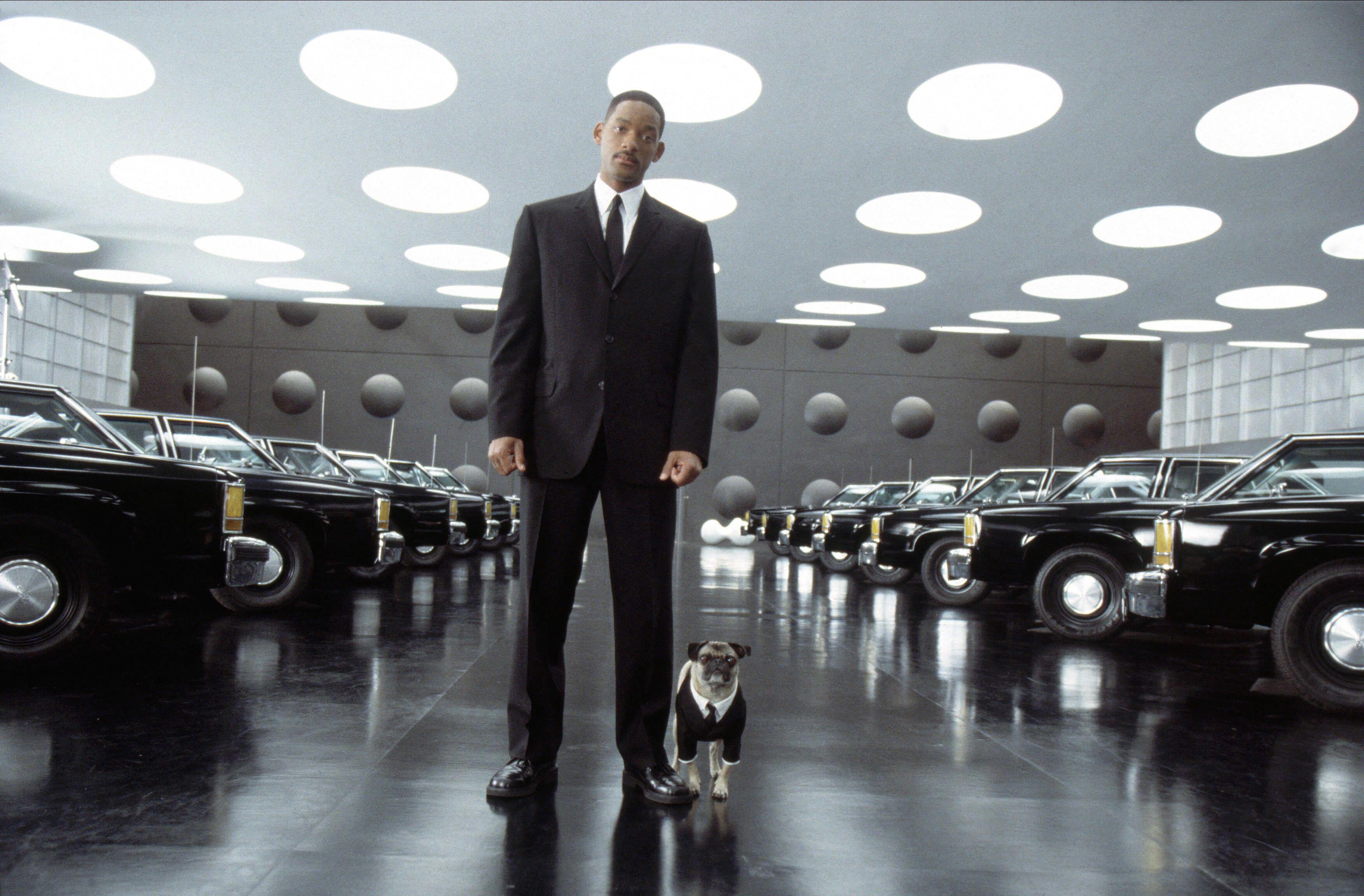 Will Smith’s Agent J stands in a futuristic showroom of black cars beside Frank the Pug, who wears a tiny black suit and tie.