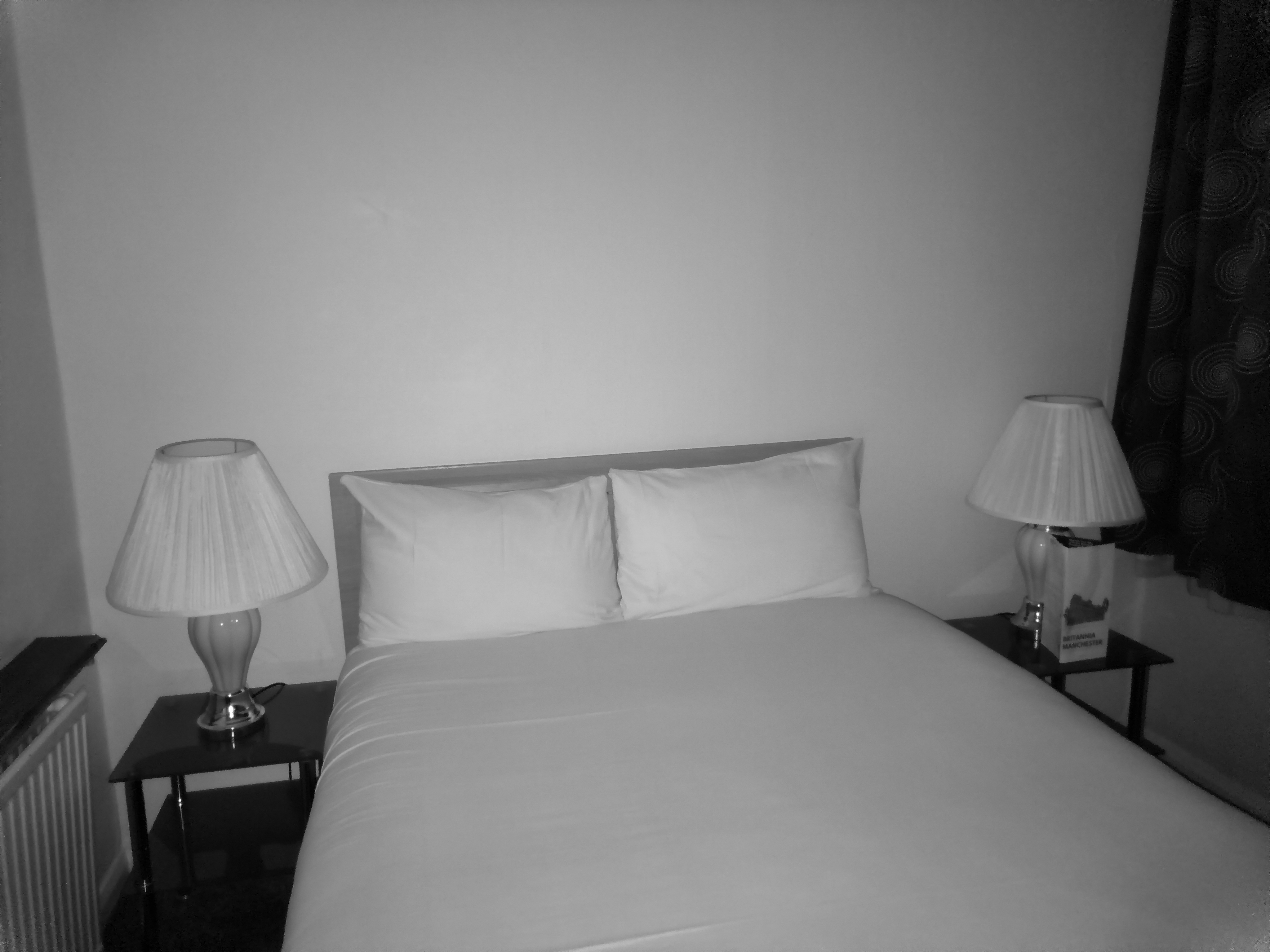 A monochrome image of a simple hotel bedroom with a neatly made double bed with white linens, matching bedside tables with lamps on either side, and dark curtains visible on the right.